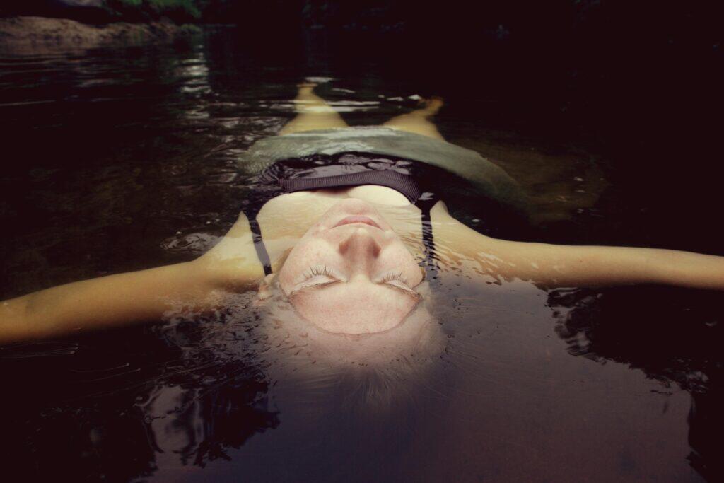 A person floating peacefully on their back in calm dark water with eyes closed, suggesting deep relaxation and weightlessness