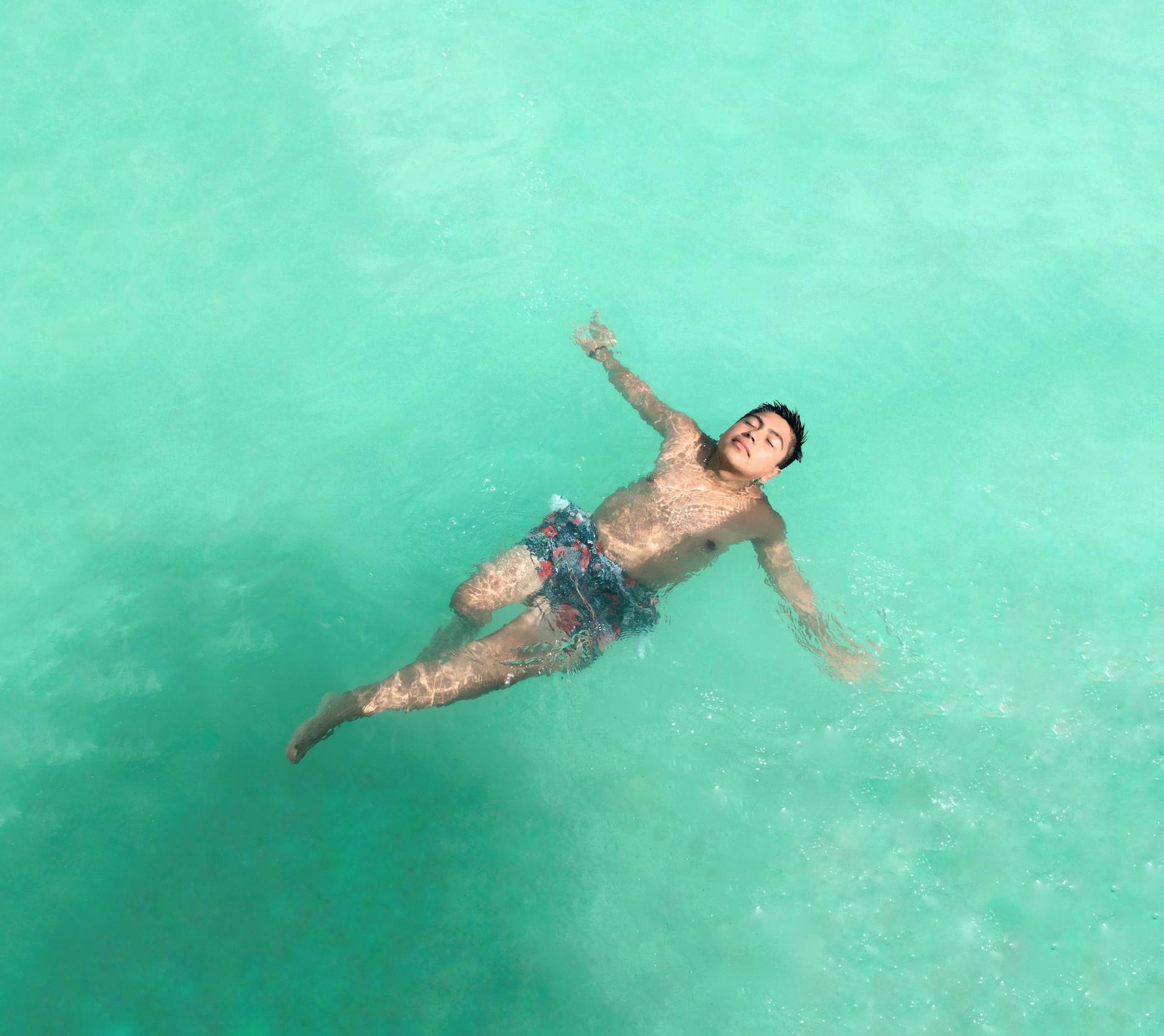 Person floating on their back in clear turquoise water seen from above, arms relaxed, eyes closed — the weightless calm of a float session