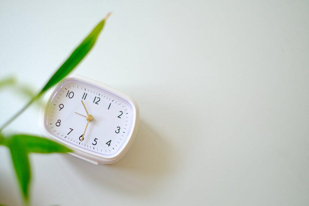 A small minimalist analogue clock beside a green plant on a calm cream background, suggesting the rhythm of regular massage