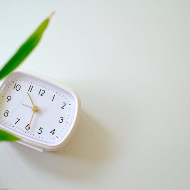 A small minimalist analogue clock beside a green plant on a calm cream background, suggesting the rhythm of regular massage