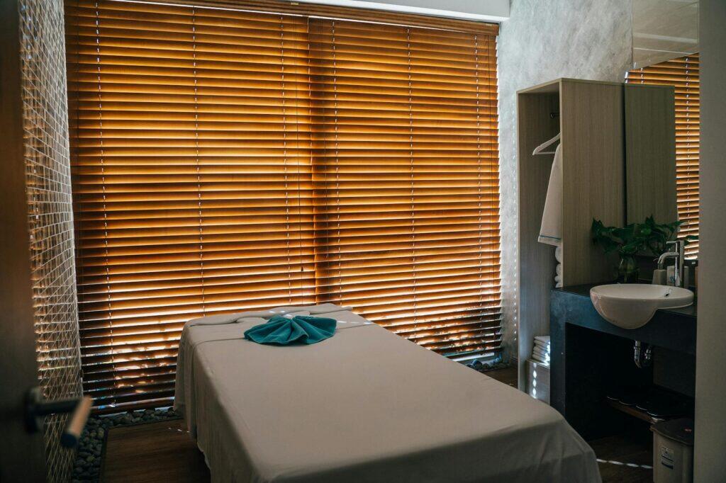 A calm massage therapy room with warm natural light through wooden blinds, neatly folded towel on a freshly prepared treatment table