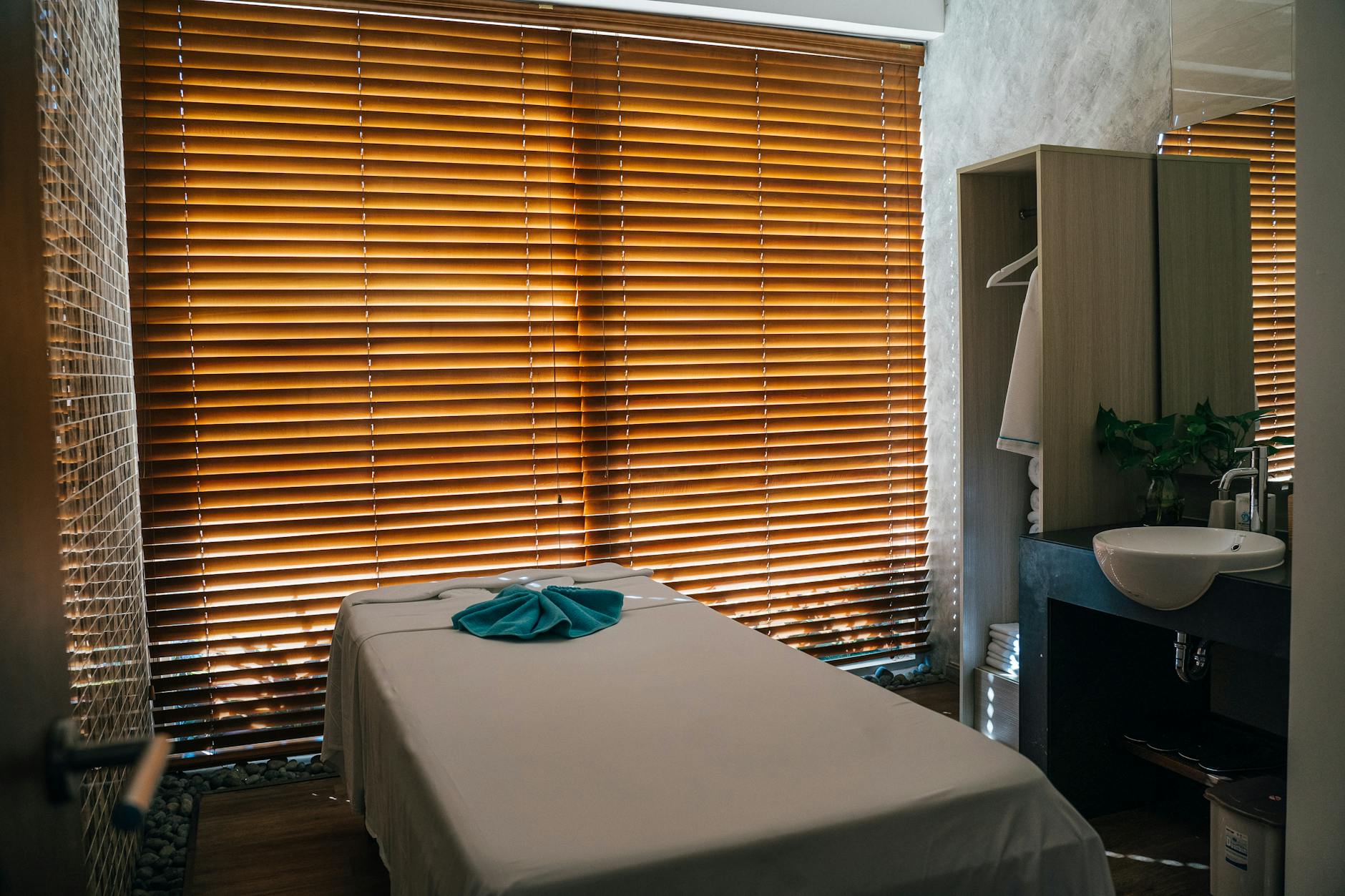 A calm massage therapy room with warm natural light through wooden blinds, neatly folded towel on a freshly prepared treatment table
