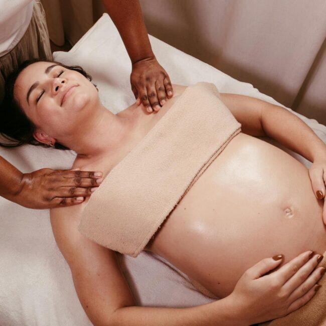 A pregnant woman receiving a calm, professional prenatal massage in a soft-lit spa setting, side-positioned and supported