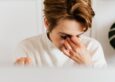 A woman in a soft white sweater rests her hand on her face at her desk, an empathetic moment of fatigue and burnout