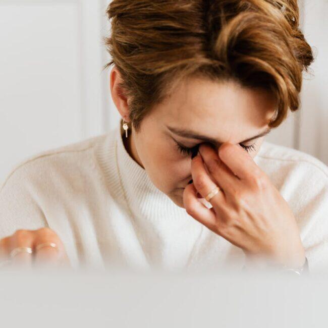 A woman in a soft white sweater rests her hand on her face at her desk, an empathetic moment of fatigue and burnout