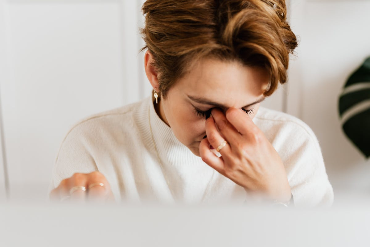 A woman in a soft white sweater rests her hand on her face at her desk, an empathetic moment of fatigue and burnout