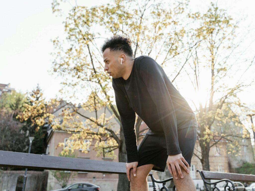 A runner pauses in a sunlit park, hands on knees, mid-recovery — the kind of moment that benefits from proper sports massage and float