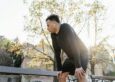 A runner pauses in a sunlit park, hands on knees, mid-recovery — the kind of moment that benefits from proper sports massage and float