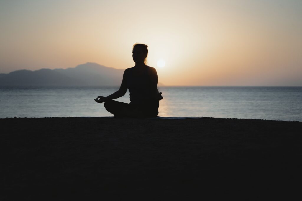 Person meditating in lotus pose silhouetted against a calm sea at sunset