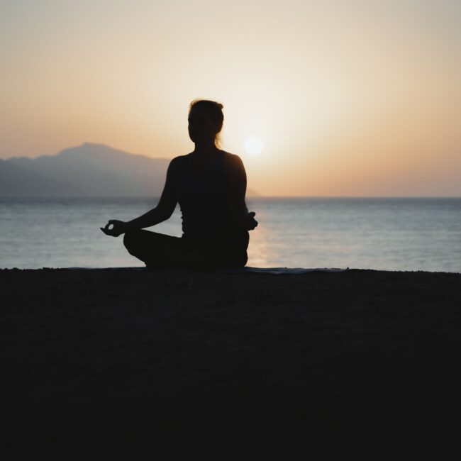 Person meditating in lotus pose silhouetted against a calm sea at sunset