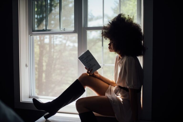 A woman sitting alone by a window with a book, looking relaxed